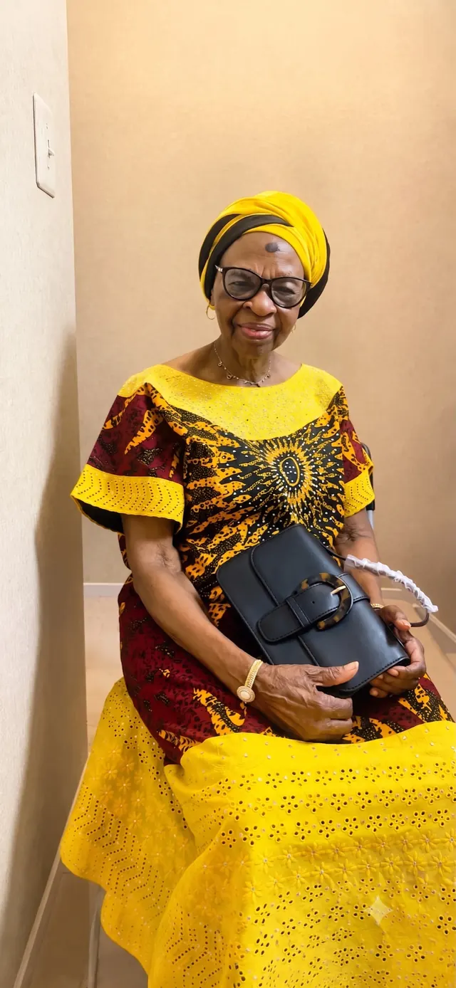 Deaconess Victoria Badejoko Lawoyin in a yellow and burgundy outfit with a yellow gele, holding a black purse.