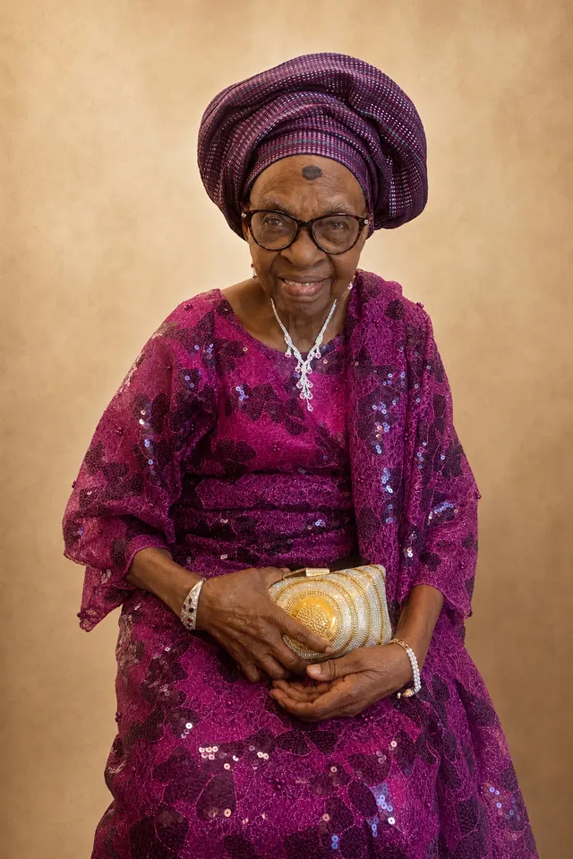 Deaconess Victoria Badejoko Lawoyin in a magenta sequined lace outfit with a purple gele, holding a gold clutch.